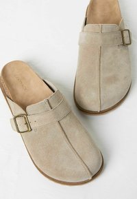 Beige Veloursleder-Clogs mit einem breiten Riemen und einer metallischen Schnalle, abgerundeter Spitze und strukturierter Oberfläche. Flache Sohle, schlichtes Design.