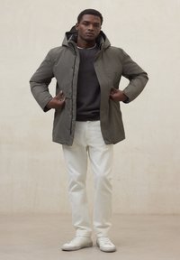 Giacca a vento imbottita verde oliva con cappuccio, dotata di chiusura con zip e bottoni a pressione. Indossata sopra un maglione grigio e abbinata a pantaloni bianchi e scarpe sportive bianche.