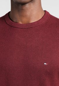 Vinröd stickad tröja med rund halsringning som har ribbad krage och mudd, med en liten logopatch i röd, vit och blå på bröstet.