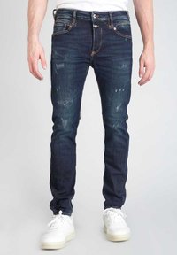 Jeans in denim skinny-fit di colore blu scuro con dettagli strappati, cuciture a contrasto e cinque tasche, abbinati a sneakers bianche.