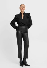 Blusa nera ondulata con colletto alto, abbinata a pantaloni neri in pelle a vita alta e stivaletti a punta. Tessuto liscio, design attillato.