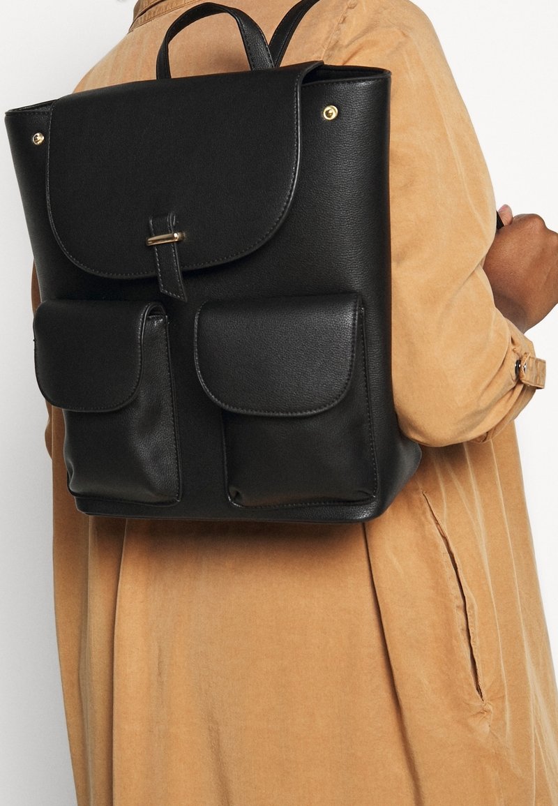 Le sac à dos en cuir noir présente un rabat de fermeture, des quincailleries dorées et deux poches avant. Texture lisse avec une forme structurée.
