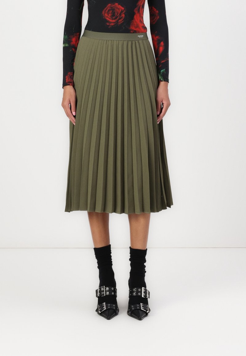 Femme portant une jupe midi plissée vert olive, un haut noir à motif de roses, des chaussettes noires et des escarpins noirs pointus avec boucles argentées.