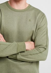 Sudadera verde oliva hecha de una suave mezcla de algodón, con un cuello redondo y puños acanalados que presentan detalles de costura visibles. Pequeño acento de logo.