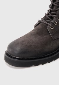 Bottine en daim gris avec bout arrondi, surface texturée, semelle en caoutchouc noire et œillets métalliques pour les lacets.