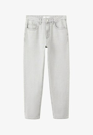 Młody mężczyzna stoi przy gładkiej białej ścianie, ubrany w kremowy sweter, jasnoszare dżinsy, brązowy pasek i brązowe buty na szarym podłożu.