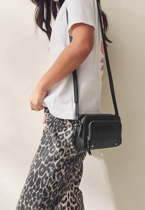 Kobieta w białej koszulce i spodniach w panterkę, niosąca małą czarną torebkę na pasku przez ramię z srebrnymi zamkami i okuciami.