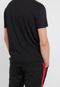 Camiseta de algodón negra de manga corta, con escote redondo, mostrada desde la parte de atrás, combinada con pantalones negros que tienen una franjas roja en el lateral.