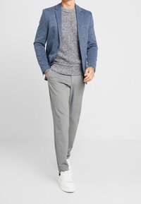 Mann, der einen blauen Blazer, einen grauen Strickpullover, helle graue Hosen und weiße Sneakers trägt, mit Händen in den Taschen vor einem einfarbigen Hintergrund.