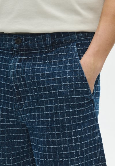 Person wearing blue checkered pants with a hand in the pocket and a white tucked-in shirt visible from the waist.