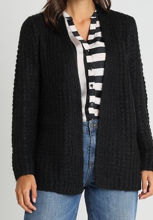 Femme portant un cardigan noir en grosse maille par-dessus une chemise à boutons rayée noir et blanc et un jean bleu.