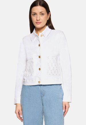 Kobieta w białej, zapinanej na guziki kurtce z koronkowymi przednimi kieszeniami i jasnoniebieskich jeansach z wysokim stanem, stojąca na jednolitym tle.