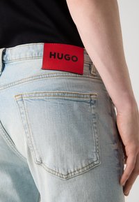 Šviesiai mėlynos džinsinės kelnės su matomais siūlais, ant juosmens esančiu raudonu odiniu ženkliuku, pažymėtu "HUGO". Matomi užpakaliniai kišenių detalės.