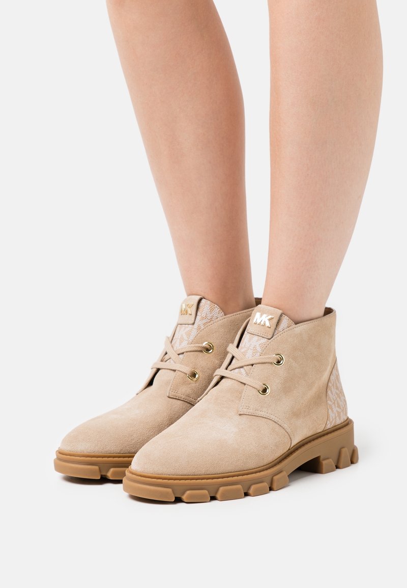 Bottines beiges en daim avec semelles épaisses en caoutchouc et œillets dorés, portées sur jambes nues, avec un panneau en tissu de marque et une étiquette avec le logo "MK".