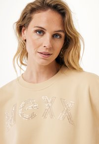 Sudadera beige con "MCXX" adornado con brillantes lentejuelas; escote redondo y textura suave. El modelo muestra un estilo casual.