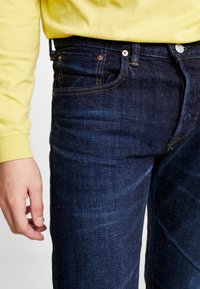 Jeans de mezclilla azul oscuro con costuras naranjas, de corte recto y diseño de cinco bolsillos. La tela tiene una ligera textura y brillo.