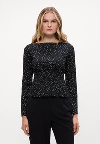 Crna majica dugih rukava s bijelim točkicama, s nabranoj strukom i peplum rubom. Glatka tekstura i dekolte u obliku čamca. Kombinirana s tamnim hlačama.