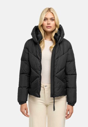 Blonde vrouw die een zwarte, gewatteerde jas met rits en hoge kraag draagt en crèmekleurige broek, staand tegen een effen achtergrond.