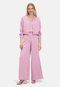 Lichtroze, satijnen twee-delige outfit met een cropped jasje en wijde broekspijpen. Voorzien van elastische manchetten en een V-hals top. Gecombineerd met tan geklede sandalen.