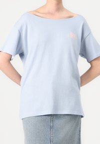 Světle modré oversized tričko vyrobené z měkké bavlny, s širokým výstřihem a krátkými rukávy. Obsahuje decentní grafický design na hrudi.