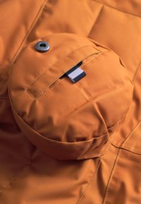 Rund orange påse med en texturerad yta, utrustad med en metallknapp och en svart-vit etikett, del av ett större vadderat tyg.