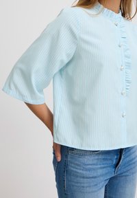 Bluză bleu deschis cu dungi verticale subțiri, guler volănițat și închidere cu nasturi. Fabricată dintr-un material neted; are mâneci trei sferturi.