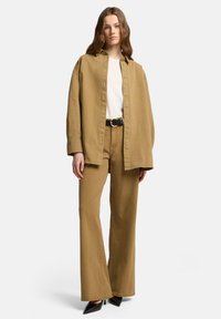Jasnobrązowa, oversize koszula na białej koszulce, w połączeniu z jasnobrązowymi rozszerzanymi spodniami i czarnymi szpilkami, prezentująca gładką fakturę materiału.
