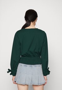 Dunkelgrünes Langarmshirt mit tailliertem Schnitt, kombiniert mit einem hellblauen, plissierten Jeansrock. Die Ärmel sind mit Schleifenakzenten versehen.