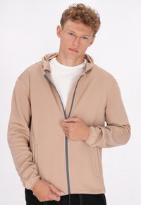 Licht beige ritshoodie, zachte stof, getailleerd ontwerp, grijze rits, twee zijzakken, elastische manchetten, gedragen over een wit shirt.