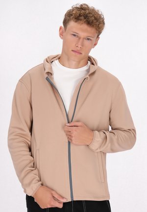 Licht beige ritshoodie, zachte stof, getailleerd ontwerp, grijze rits, twee zijzakken, elastische manchetten, gedragen over een wit shirt.