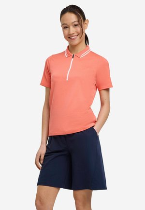 Lächelnde Frau mit dunklen Haaren im Pferdeschwanz trägt korallenfarbenes Poloshirt mit kurzen Ärmeln und weißem Reißverschluss sowie marineblaue Shorts bis zum Knie, steht mit der Hand in der Tasche.