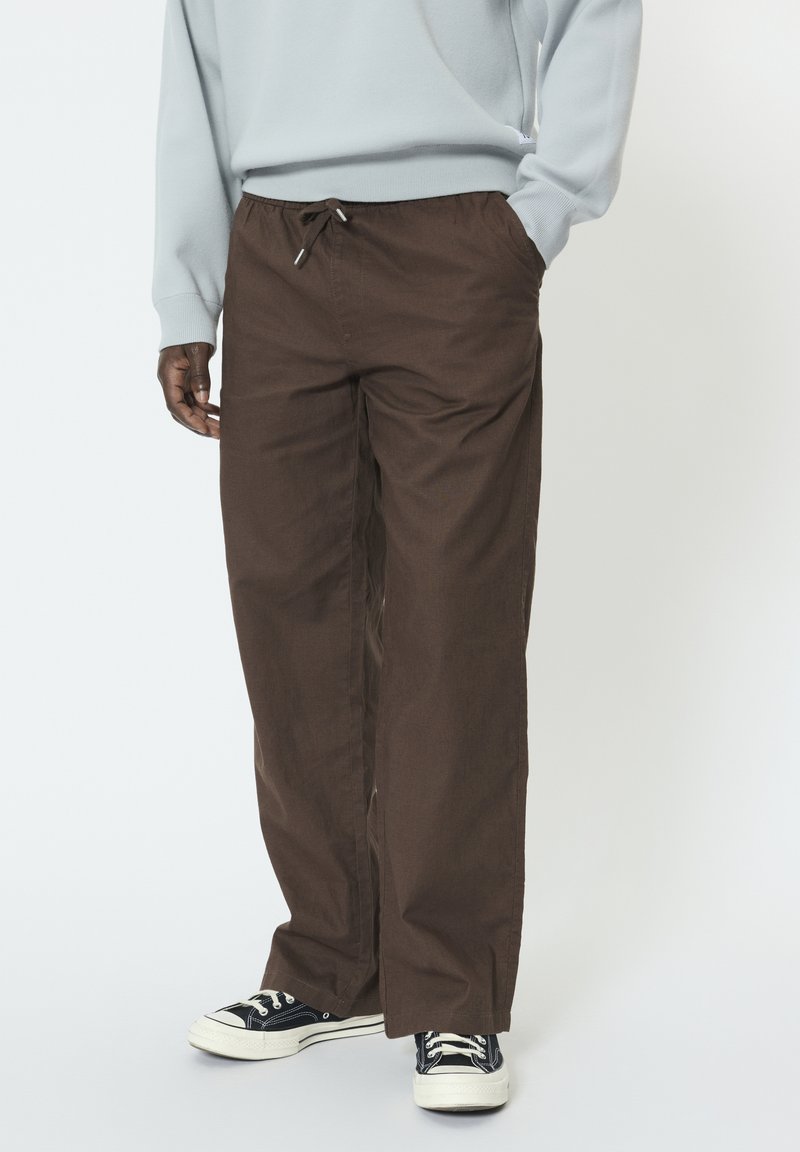 Mustanahaline inimene, kes kannab pruunide paelaga pükse, helehallit kampsunit ja must-valgeid tenniseid, seisab ühe käega taskus.