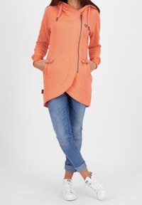 Veste à capuche pêche avec zip, poches latérales et ourlet arrondi. Tissu doux et détails de couture contrastants. Associée à un denim et des baskets.