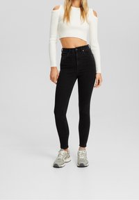 Jeans skinny negros de cintura alta, combinados con una camiseta corta blanca de canalé con hombros al descubierto. La modelo lleva zapatillas grises. Diseño minimalista.