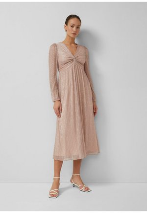 Kobieta stojąca w błyszczącej różowej sukience o długości midi z długim rękawem i przednim splotem, w połączeniu z białymi sandałami na niskim obcasie z paskami.