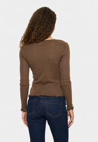 Vrouw met krullend haar, gekleed in een aansluitend bruin langekatoenen shirt en donkerblauwe jeans, staande met haar rug naar de camera tegen een effen achtergrond.
