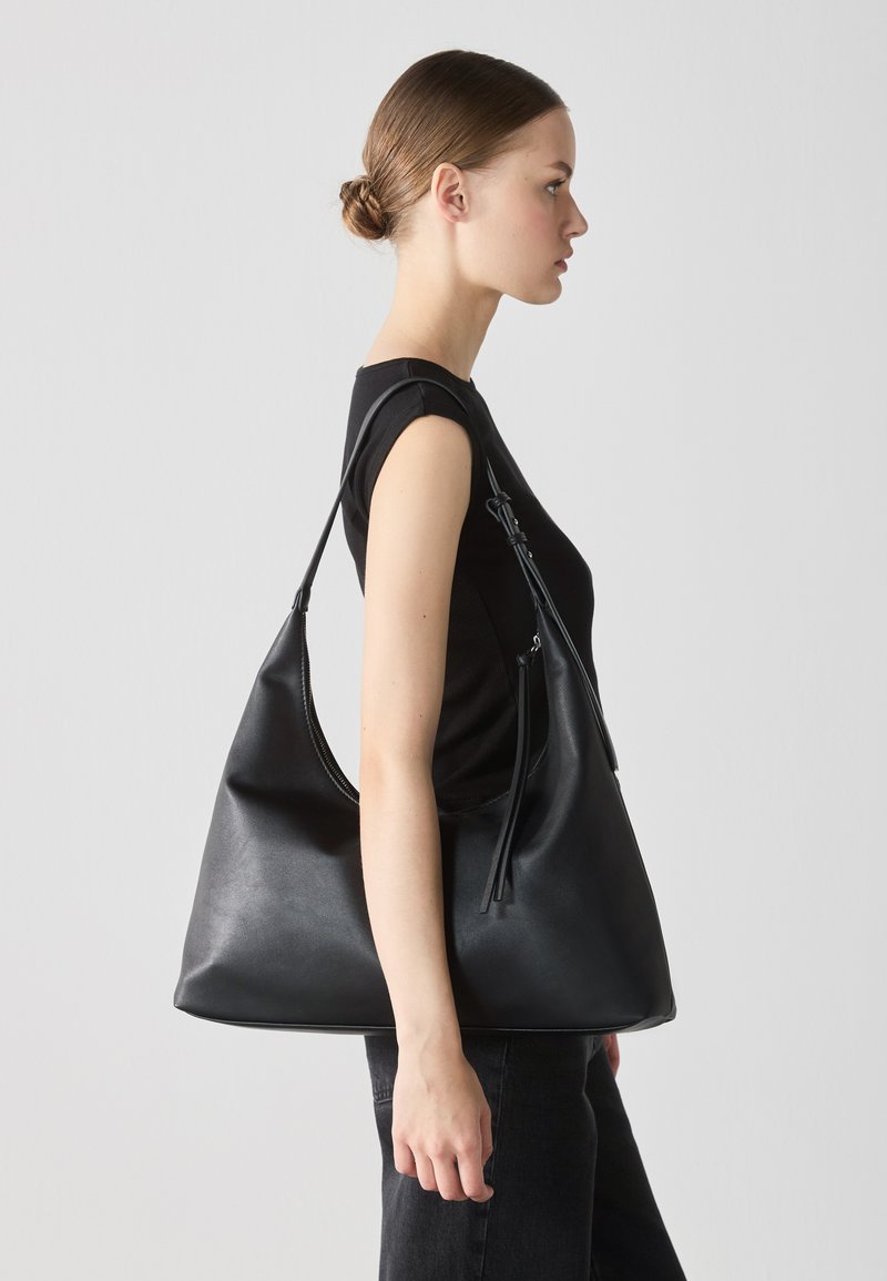 Bolso hobo de cuero negro con una silueta curvada, correa de hombro ajustable y un sutil detalle de cremallera; textura suave y diseño minimalista.