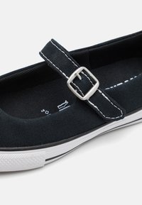 Chaussure en toile noire avec une bride ornée d'une boucle argentée. Coutures blanches en accent et semelle en caoutchouc avec des rayures noires et blanches.