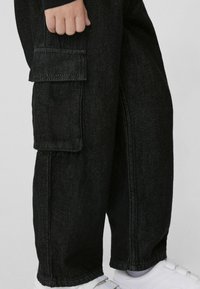 Czarne dżinsowe spodnie cargo z boczną kieszenią, w luźnym kroju, oraz białe trampki. Materiał ma lekko teksturowany wygląd.