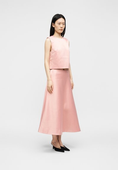 Γυναίκα φοράει ροζ crop top χωρίς μανίκια και αντίστοιχη midi φούστα με ψηλή μέση, με μαύρες μυτερές ψηλοτάκουνες χαμηλές γόβες, στέκεται μπροστά σε απλό φόντο.