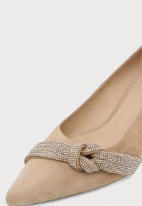 Chaussure plate en daim beige à bout pointu avec une bride en strass scintillante nouée en nœud sur le devant.