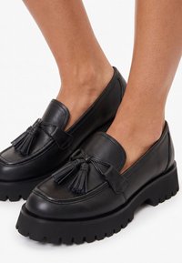 Mocasines de cuero negro con borlas y suelas gruesas con tacos, usados con los pies desnudos, mostrados sobre un fondo blanco liso.