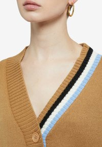 Donna che indossa un cardigan beige con bordi a righe blu, bianche e nere e un orecchino a cerchio dorato intrecciato visibile sull'orecchio destro.