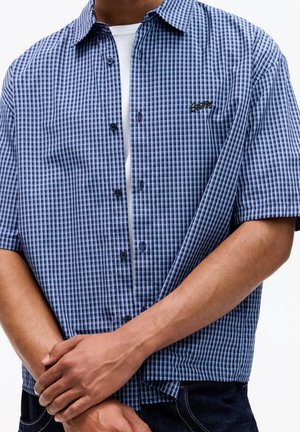 Homme portant une chemise à manches courtes à carreaux bleus et noirs par-dessus un T-shirt blanc, les mains croisées à la taille.