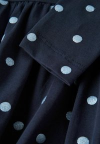 Granatowy materiał z srebrnymi kropkami; zebrany design o miękkiej teksturze; zbliżenie pokazuje szew rękawa oraz szczegóły wzoru w kropki.