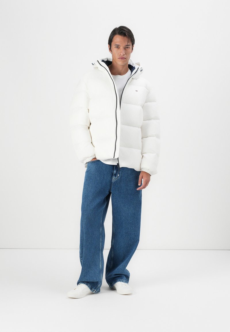 Witte pufferjack met een ritssluiting, verstelbare capuchon en marineblauwe voering; gedragen over een wit shirt en gecombineerd met losse blauwe spijkerbroeken en witte schoenen.