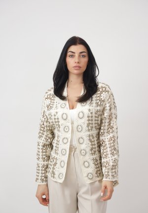 Vrouw met lang donker haar, die een beige gebreid vest met een haakpatroon draagt over een wit topje en lichtgekleurde hoge taillebroek, staande tegen een effen achtergrond.
