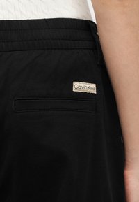 Πλευρική όψη ατόμου που φοράει μαύρο παντελόνι με μικρή πίσω τσέπη και λευκή ετικέτα Calvin Klein πάνω από αυτήν.