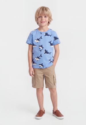Blond dreng smilende, iført en blå t-shirt med hajtryk, beige cargoshorts og brune slip-on sko, står med den ene hånd i lommen.