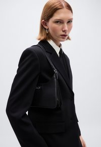 Μαύρη δερμάτινη τσάντα ώμου με καμπυλωτό σχήμα, διακριτική υφή και ασημένια μεταλλικά στοιχεία, συνδυασμένη με μαύρο κοστούμι και λευκό πουκάμισο.